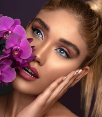 A beautiful blonde woman holds a purple orchid, showcasing the elegance of Kaya by Reema Salon's facial services.